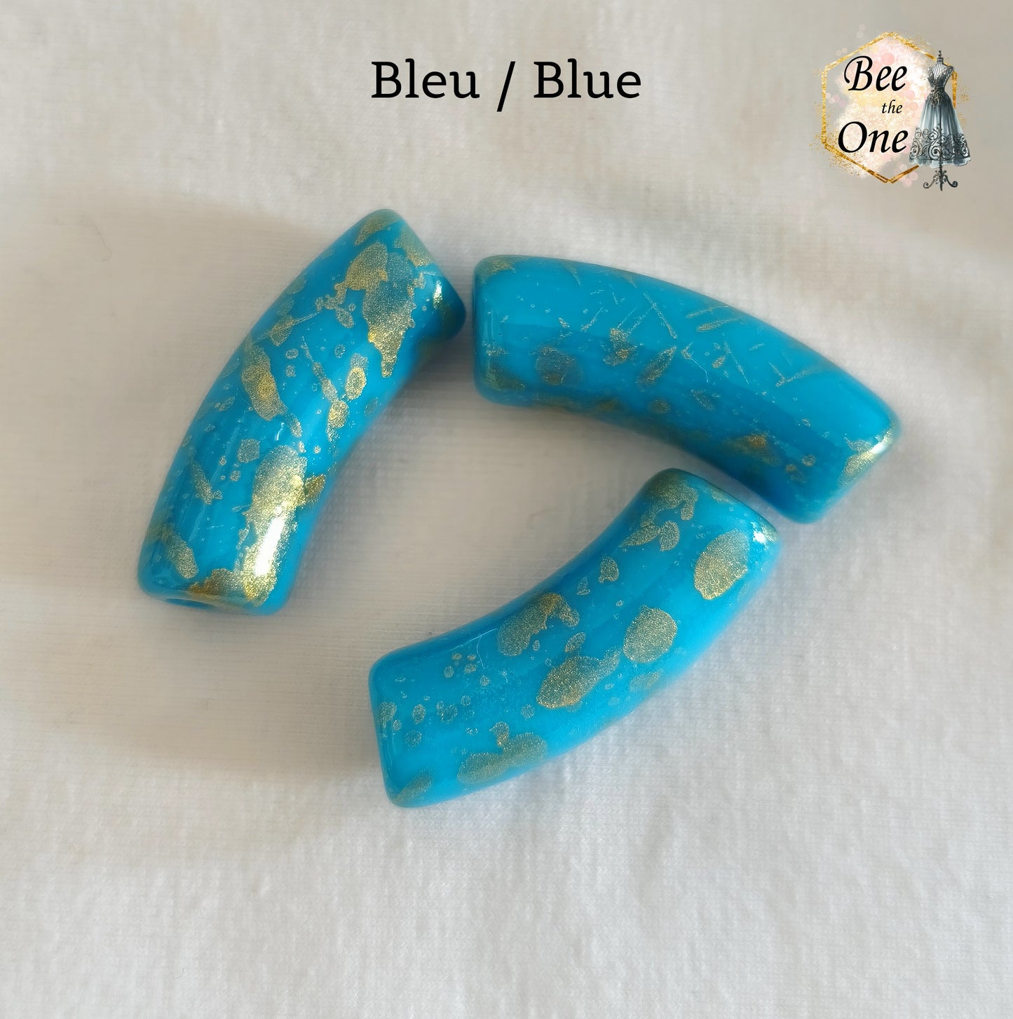 Grandes perles tubes incurvées en acrylique mesurant 34x11 mm. Les perles sont couleur bleu et ornées d'éclats et de mouchetures dorées, disposées sur un fond blanc.