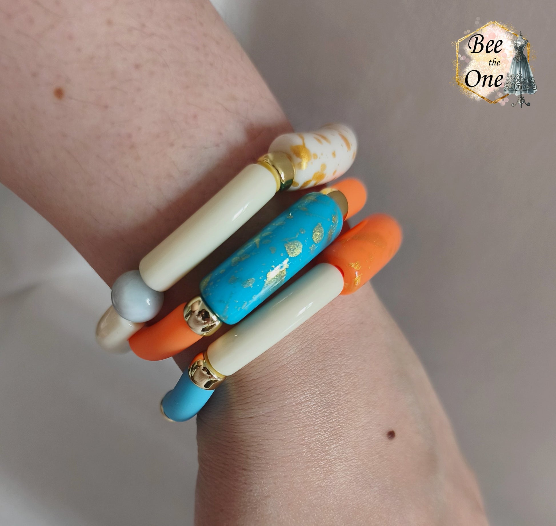 Set de trois bracelets élastiques faits main pour adulte, composés de perles tubes incurvées en acrylique aux teintes bleu azur, orange vif et crème, avec détails marbrés dorés et séparateurs dorés, présentés sur un poignet de femme