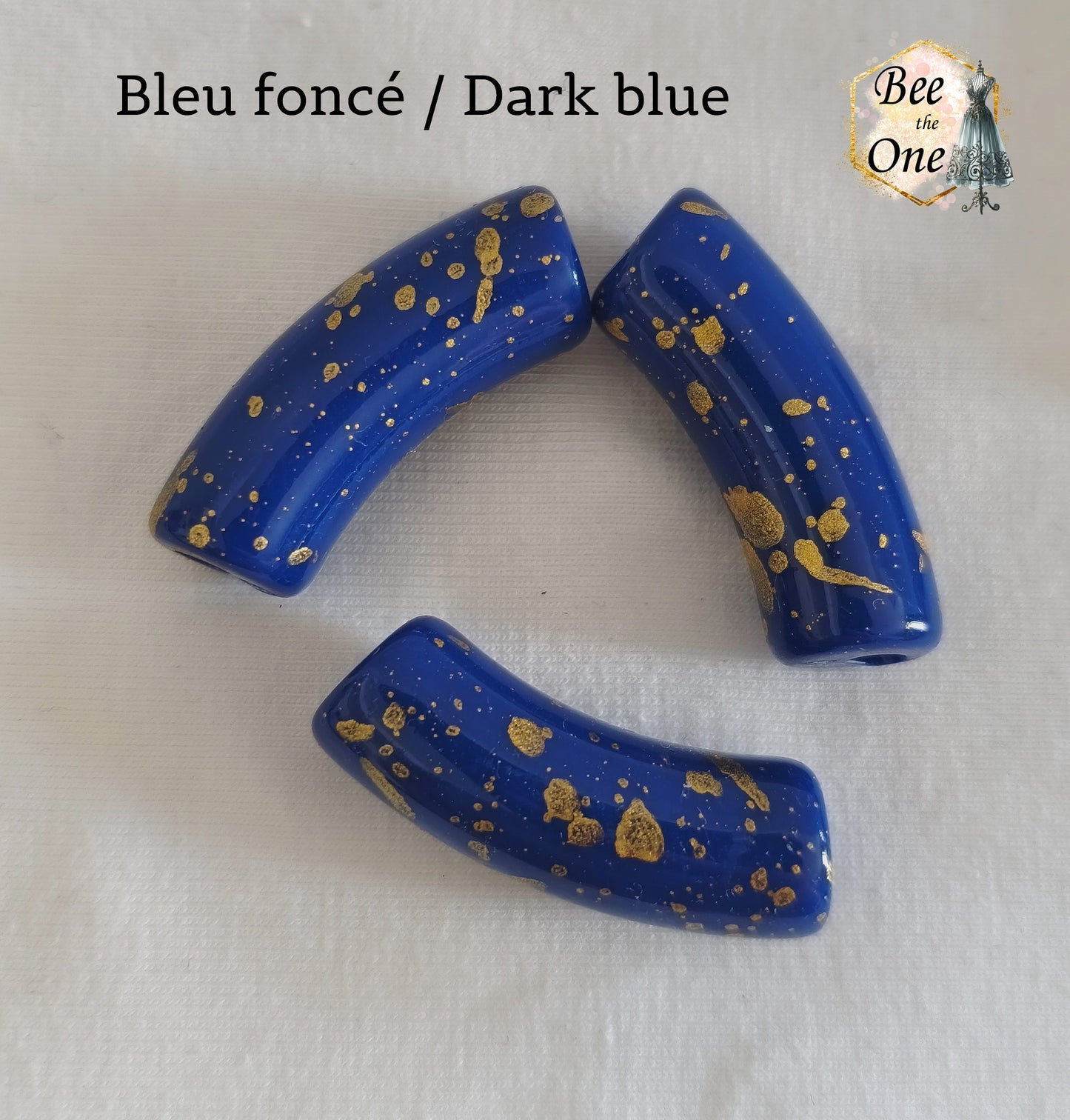 Grandes perles tubes incurvées en acrylique mesurant 34x11 mm. Les perles sont couleur bleu foncé et ornées d'éclats et de mouchetures dorées, disposées sur un fond blanc.