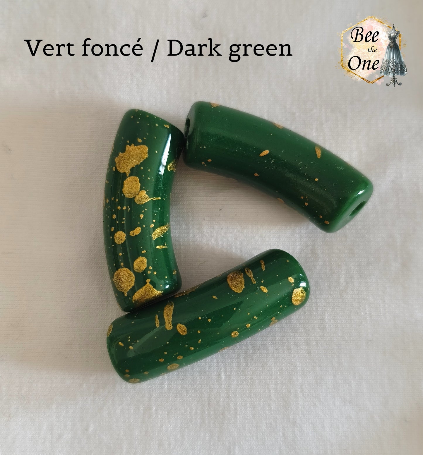 Grandes perles tubes incurvées en acrylique mesurant 34x11 mm. Les perles sont couleur vert foncé et ornées d'éclats et de mouchetures dorées, disposées sur un fond blanc.