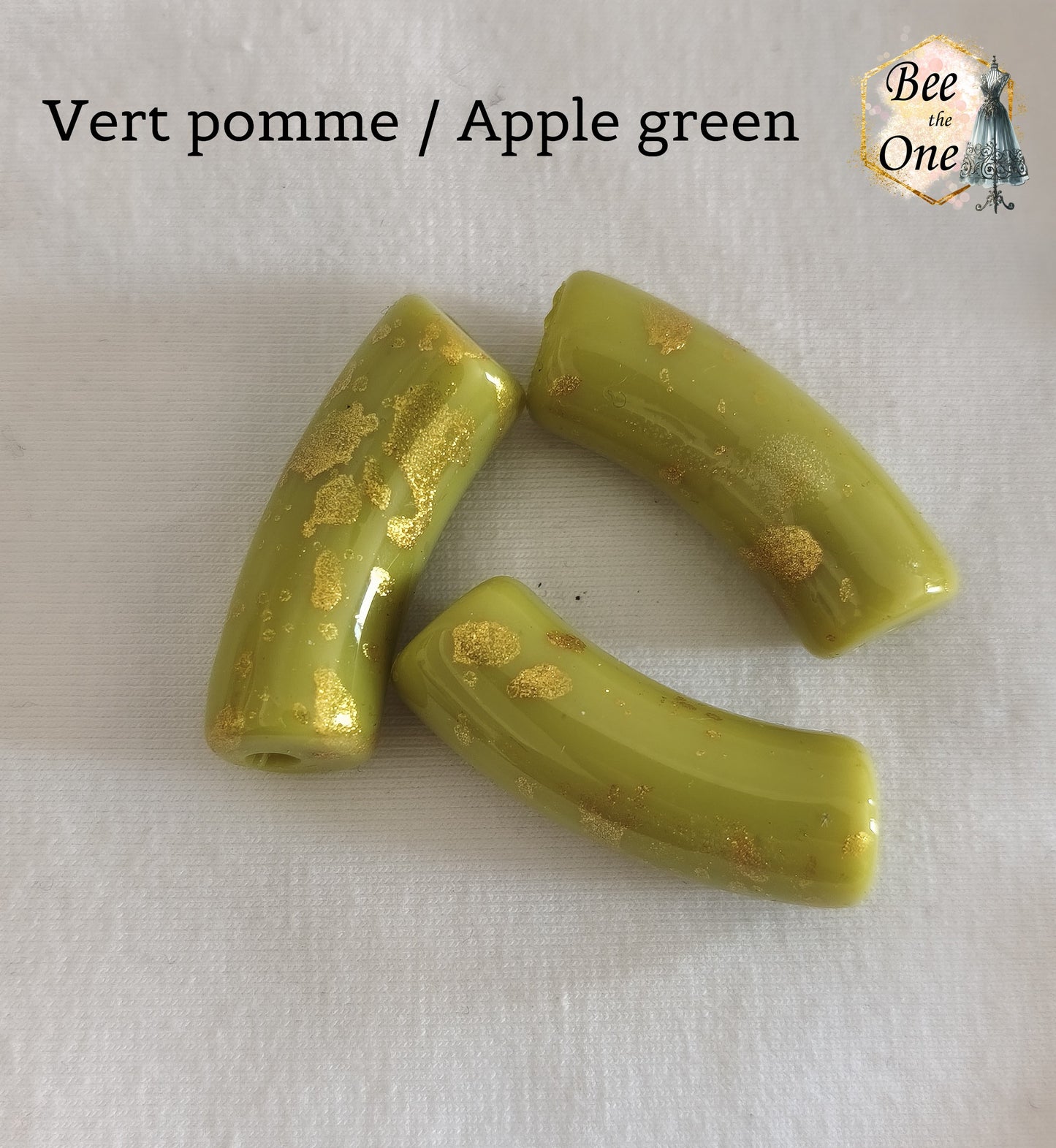 Grandes perles tubes incurvées en acrylique mesurant 34x11 mm. Les perles sont couleur vert pomme et ornées d'éclats et de mouchetures dorées, disposées sur un fond blanc.